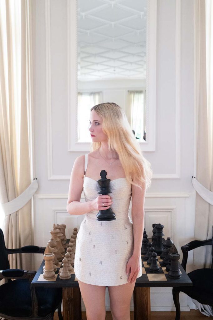 Braut in einem weißen Cocktailkleid blickt nach rechts und hält eine schwarze Schachfigur in den historischen Innenräumen von Hotel Weissenhaus.